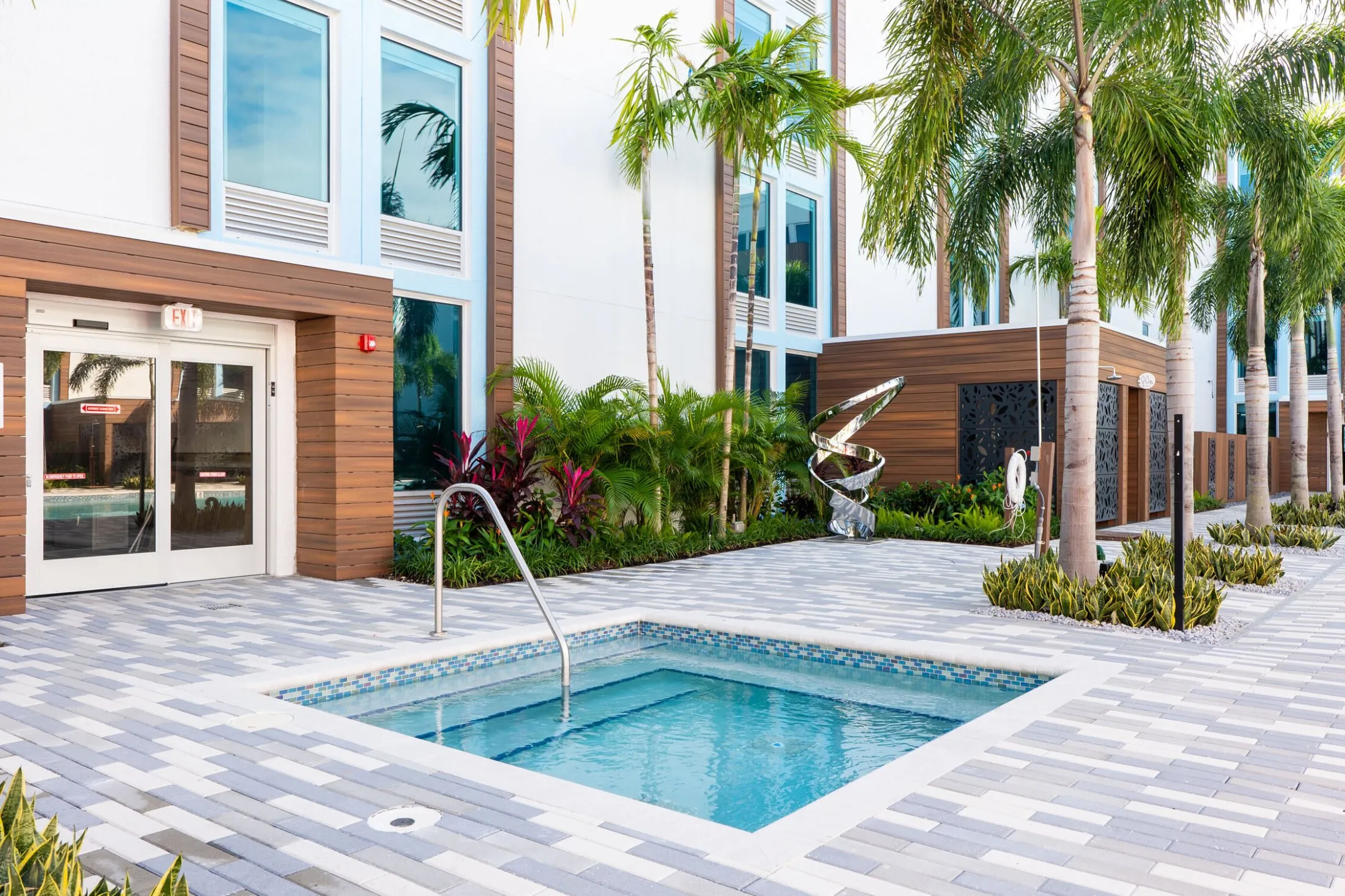 A small pool with a metal handrail, surrounded by palm trees and a building with brown and white walls.